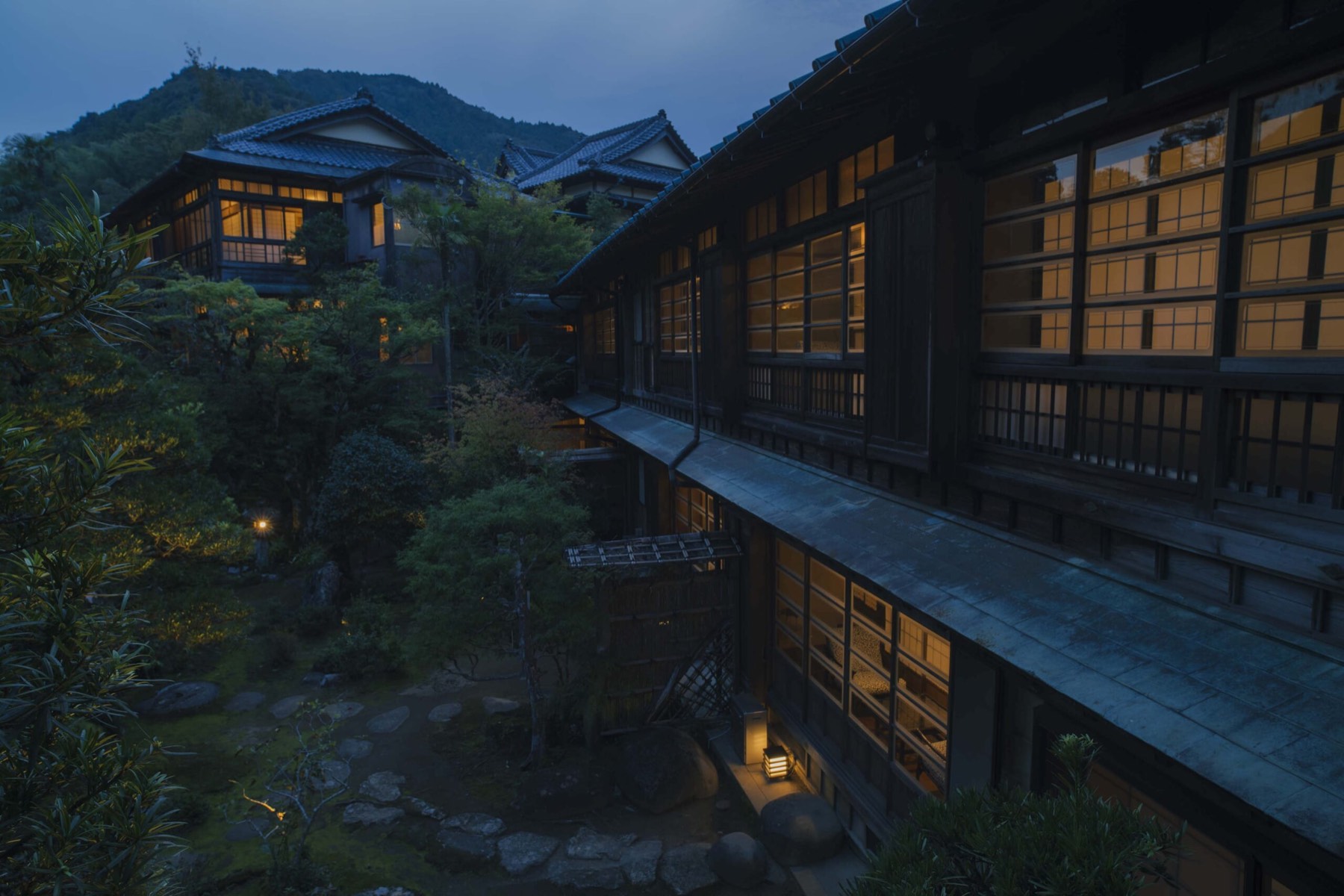 The wooden ryokan at dusk, windows lit, mountain behind